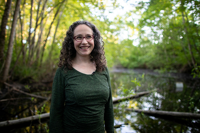 A woman with curly brown hair and glasses smiles while standing in front of a pond surrounded by green trees. She is wearing a long-sleeved green shirt, and the background is softly lit by natural light.