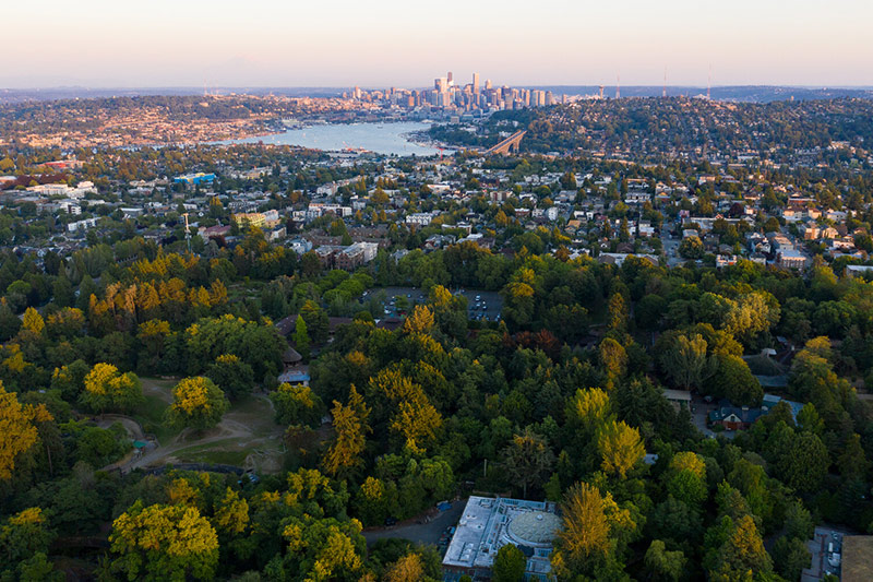 Aerial view of a cityscape with dense green trees and the zoo in the foreground, residential neighborhoods in the middle, and a downtown skyline in the background under a clear sky.