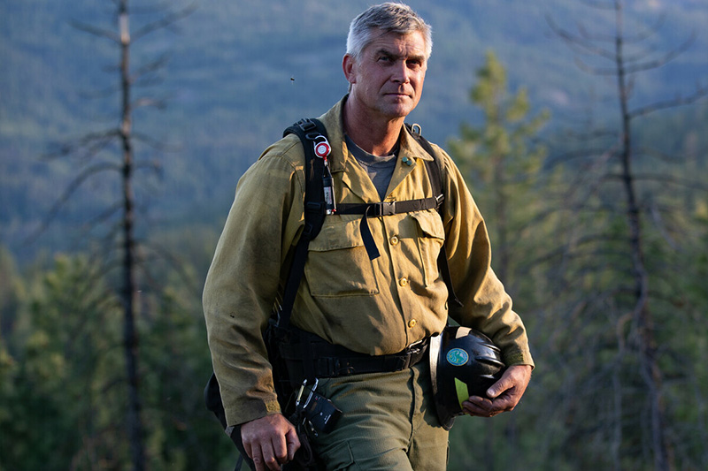 A firefighter in yellow protective gear stands outdoors, holding a helmet with one hand. Trees and hills are visible in the blurred background. The lighting suggests early morning or late afternoon.