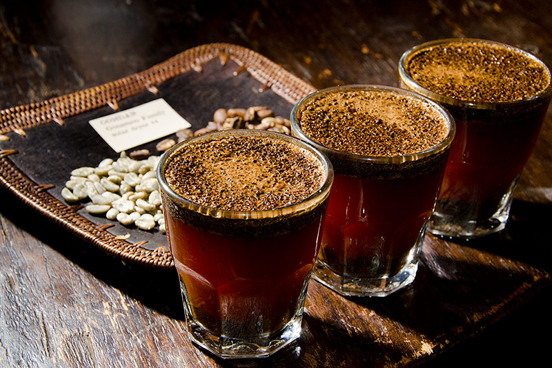 Three glasses of freshly brewed coffee with grounds floating on top are lined up beside a tray holding green and roasted beans.