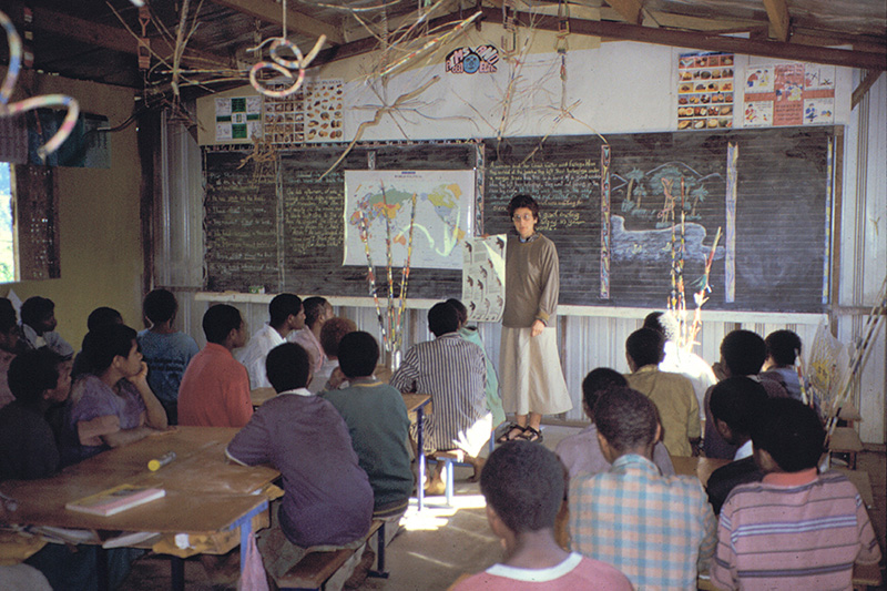 A teacher stands at the front of a classroom, pointing to a world map on a blackboard as students listen attentively amid handwritten notes and educational posters.