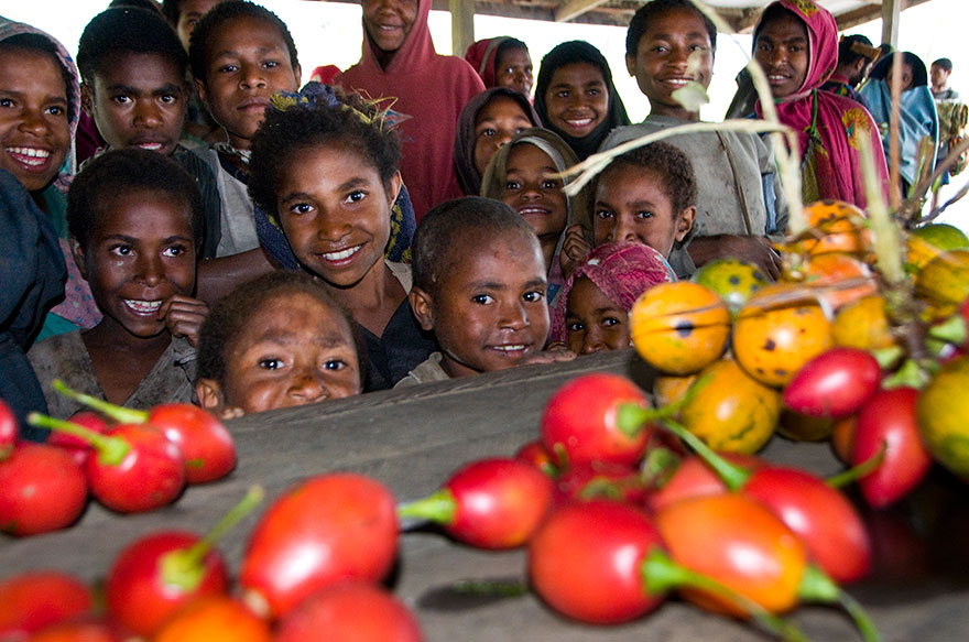 A group of children and adults smile and stand together behind a table covered with bright orange and yellow fruits in a market setting with a wooden roof overhead.