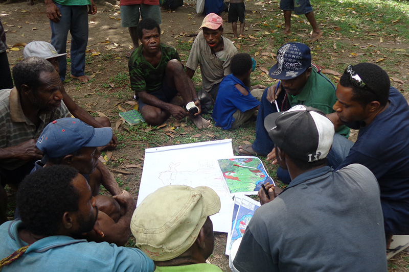 A group of men sit on the ground outdoors, closely gathered around maps and papers discussing. Trees and leaves surround them as they focus on conservation efforts.