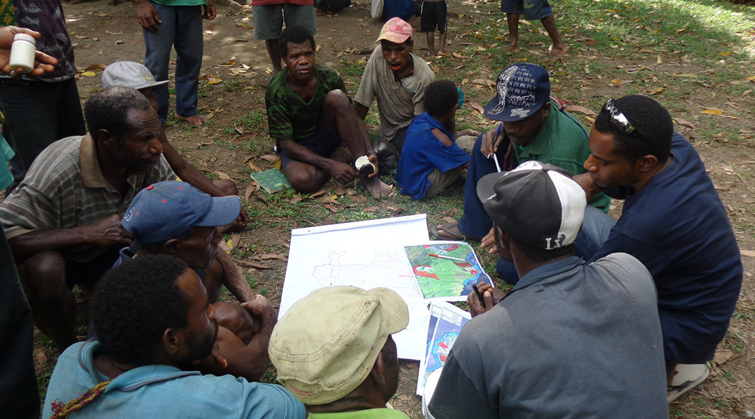 A group of people sit outdoors in a circle, looking at maps and papers spread on the ground. Most are in casual clothing and hats, surrounded by greenery and scattered leaves.