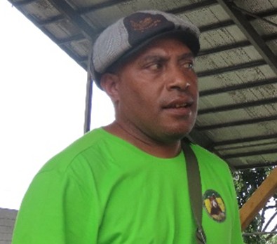 Desmond Nalong. A man wearing a gray flat cap and a bright green shirt stands under a metal roof, looking slightly to the side.