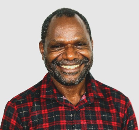 James Jio. A man with short curly hair and a beard, wearing a red and black plaid shirt, smiles warmly at the camera against a plain light background.
