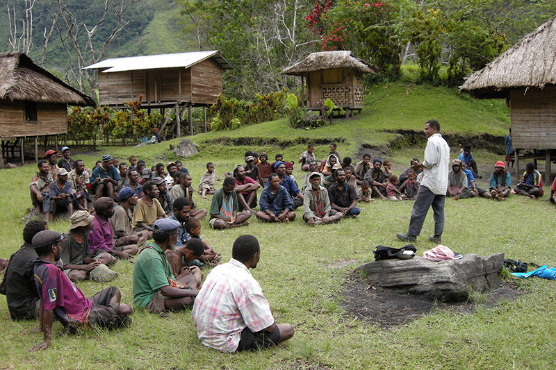 A group of people sit on grass in front of wooden huts, listening to a man standing and speaking outdoors in a rural, lush green village setting.