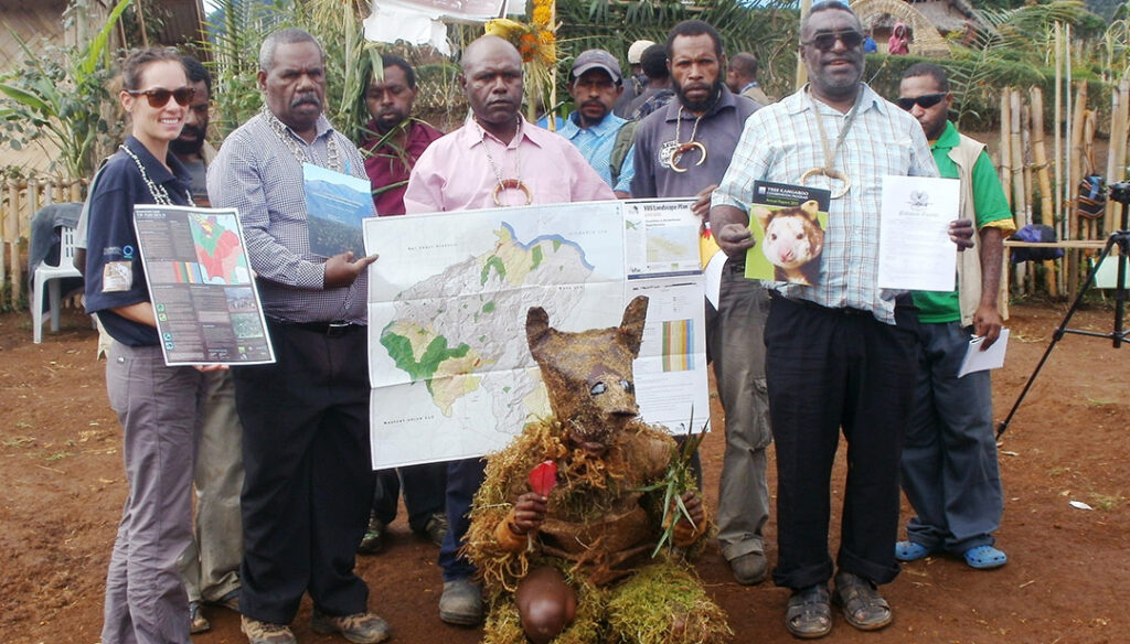 A group of people outdoors, some holding maps and documents. One person kneels in front in a leafy costume and mask, with trees and a bamboo fence in the background.