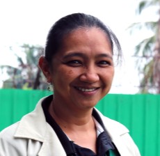 Roebeck Bacani. A woman with dark hair tied back smiles at the camera. She is wearing a light-colored jacket over a black shirt, with a green fence and palm trees in the blurred background.