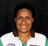 Sharon Erick. A woman with short curly hair smiles at the camera. They are wearing a white shirt with green sleeves and a collar, standing in front of a plain black background.