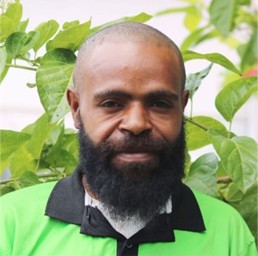 Teddy Hesing. A man with a full beard and short hair wearing a green collared shirt stands in front of leafy green plants, smiling slightly at the camera.