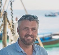 Trevor Holbrook, a man with short gray hair and a beard, wearing sunglasses on his head and a light blue shirt, smiles outdoors with boats and water blurred in the background.