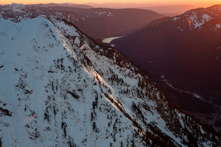 A snowy mountain ridge is bathed in warm sunset light, with forested hills and a distant lake in the background. The sky glows in soft orange tones, highlighting the contrast between snow and shadow.