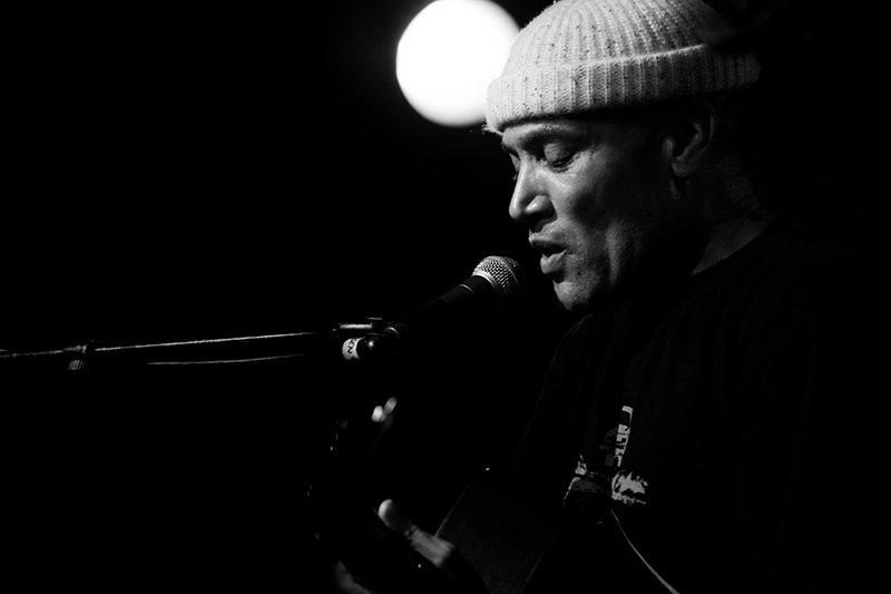 Black-and-white photo of a man in a knit hat singing into a microphone onstage, with a strong light glowing behind him, creating a dramatic and intimate atmosphere.