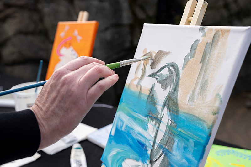 A hand holding a paintbrush works on a canvas featuring a penguin against a blue and brown background; another colorful painting is visible in the background.