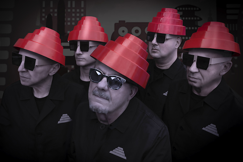 Five people wearing matching black outfits, red energy dome hats, and black square sunglasses stand together, facing various directions. The background is dark with abstract shapes and patterns.