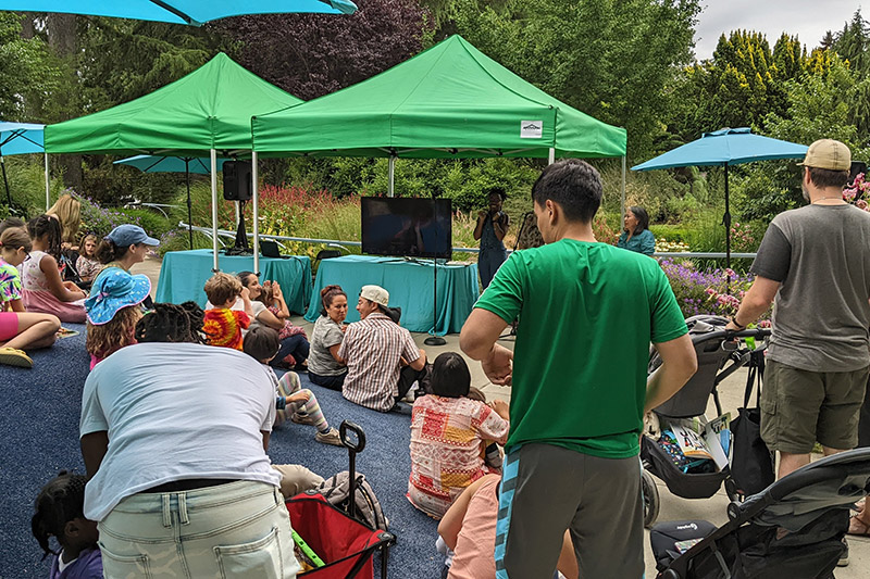A group of adults and children gather outdoors under green canopy tents, sitting and standing around a TV screen. Blue umbrellas and lush greenery surround the area, creating a lively, relaxed atmosphere.