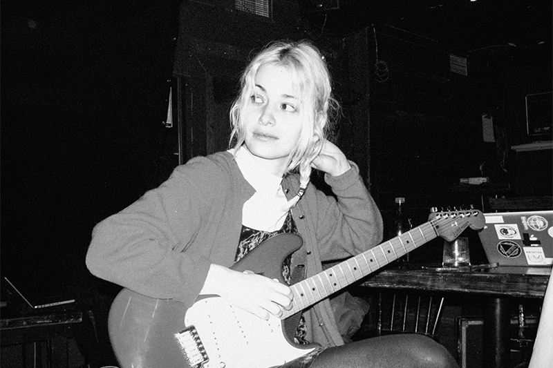 A woman with light hair sits at a table in a dimly lit room, holding an electric guitar on her lap and looking off to the side. There is a laptop with stickers and a glass of water on the table behind her.