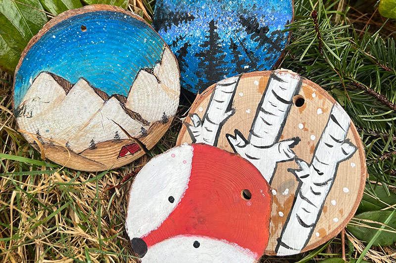 Four round wooden ornaments painted with different nature scenes: snowy mountains and a red cabin, a starry night sky with pine trees, white birch trees with snow, and a red fox face. All are lying on grass and fir branches.