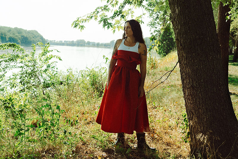 A person in a red dress and white top stands by a tree, looking to the side. They are outdoors near a lake with green foliage and grass, and sunlight filters through the branches.