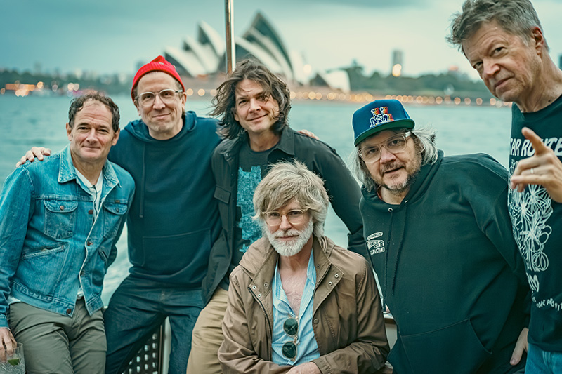 Six men pose together outdoors with the Sydney Opera House in the background across the water. They are casually dressed and smiling, suggesting a relaxed and friendly atmosphere.