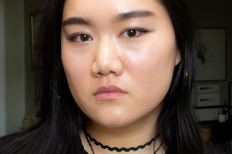 A person with long black hair, wearing hoop earrings and a black choker necklace, looks directly at the camera with a neutral expression. The background is indoors with blurred furniture.