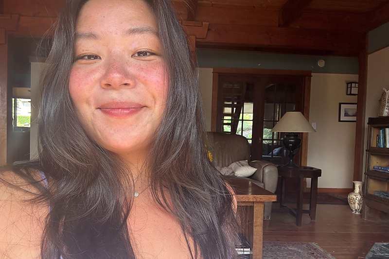 A woman with long brown hair and a natural, slightly flushed face smiles at the camera inside a cozy wooden room with bookshelves, a lamp, and a large armchair in the background.