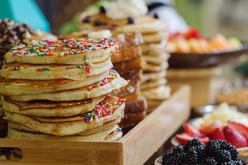 A stack of fluffy pancakes topped with colorful sprinkles and syrup sits on a wooden tray, surrounded by various fruits including strawberries and blackberries.