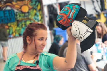 A woman with red hair holds up a trucker hat featuring a colorful octopus design, surrounded by people working at tables in a lively, art-filled space.