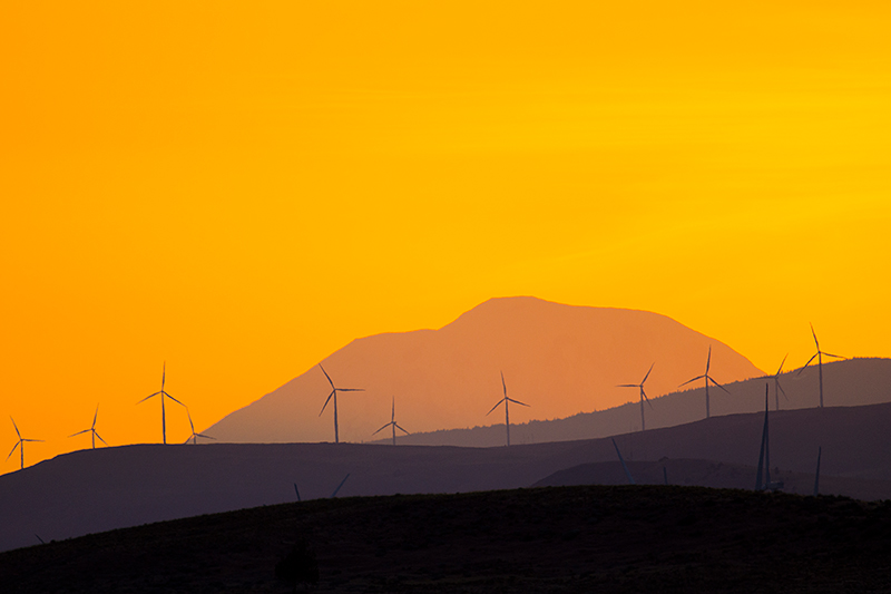 Silhouettes of wind turbines on rolling hills with a mountain in the background, set against a bright orange and yellow sunset sky.