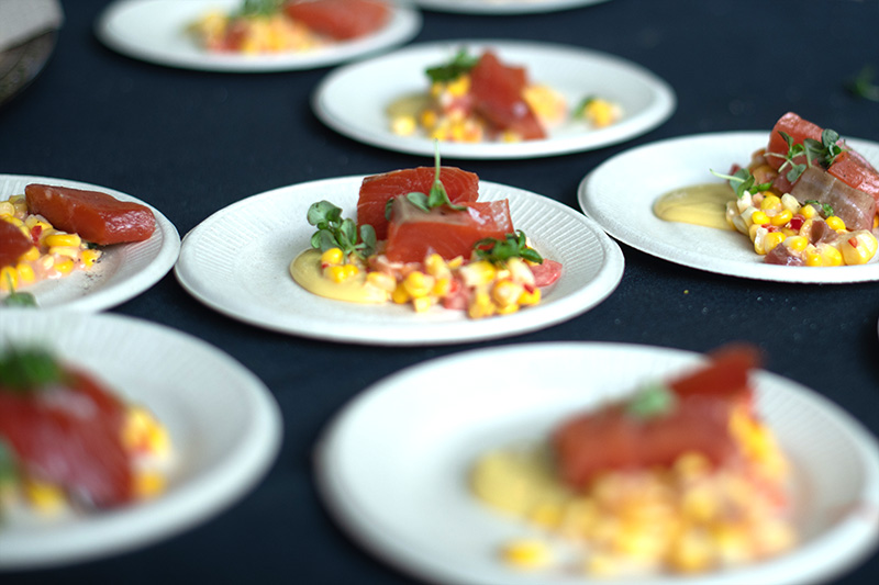 Several white plates with a gourmet dish featuring slices of cured fish, a corn salad, microgreens, and a light yellow sauce, arranged on a dark tablecloth.