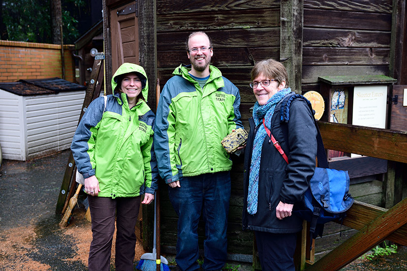 Three adults stand outside near a wooden structure, smiling at the camera. Two wear green zoo staff jackets. The third person wears a blue scarf and black coat. The ground is wet, suggesting recent rain.