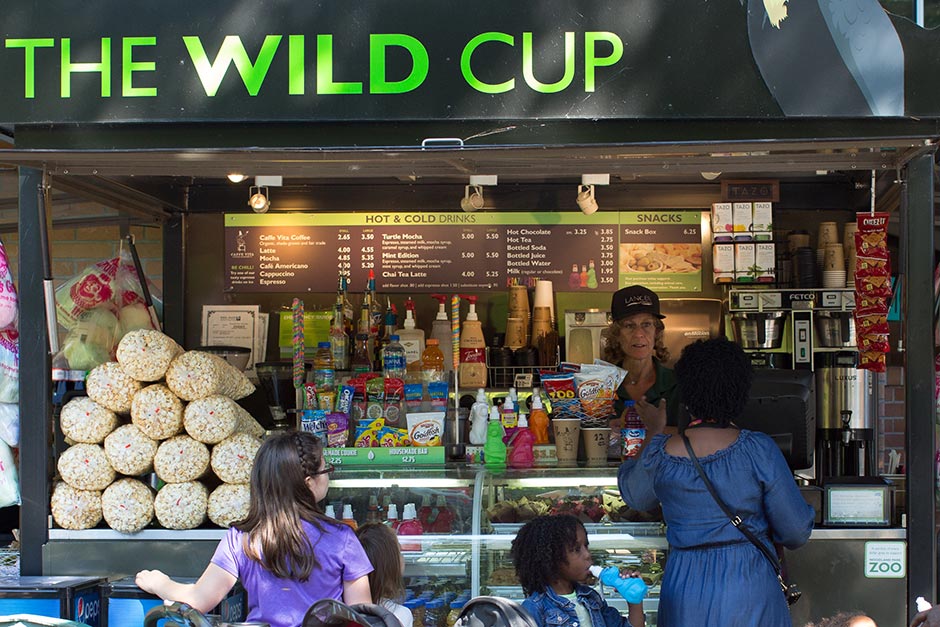 A snack stand called The Wild Cup displays popcorn, chips, drinks, and snacks. Two women and two children stand at the counter while a staff member assists them. The menu is visible behind the counter.