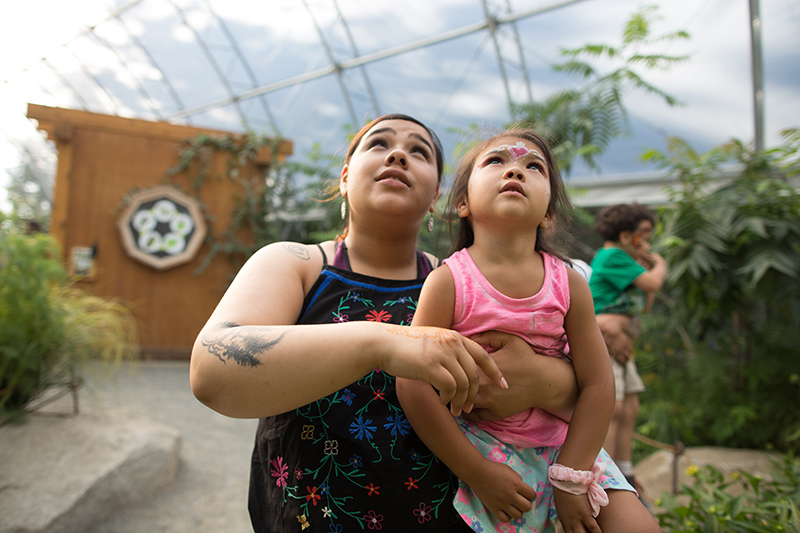 A woman holding a young girl looks upward in a greenhouse, both appearing curious. The girl has face paint and wears a pink outfit. Plants and another child are visible in the background.