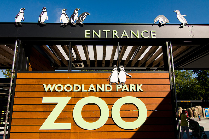 The entrance to Woodland Park Zoo with large signs and cartoon penguin decorations on top of the structure against a clear blue sky.