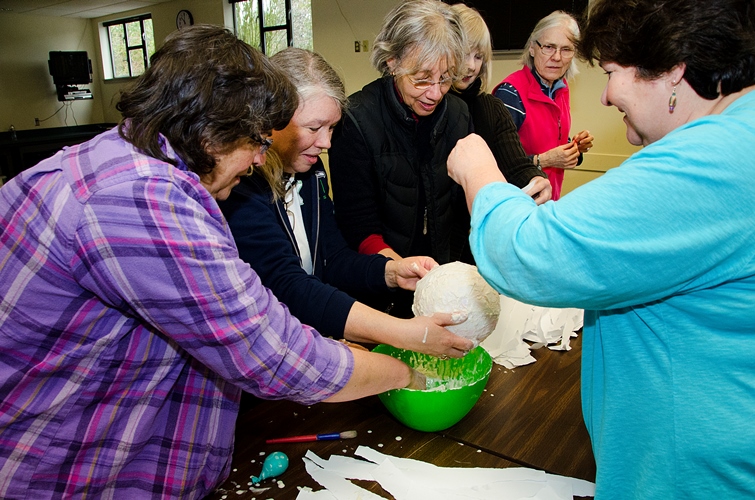 A group of women gather around a table, working together on a craft project with a papier-mâché balloon over a green bowl. Torn white paper and glue are spread around as they smile and concentrate on the activity.