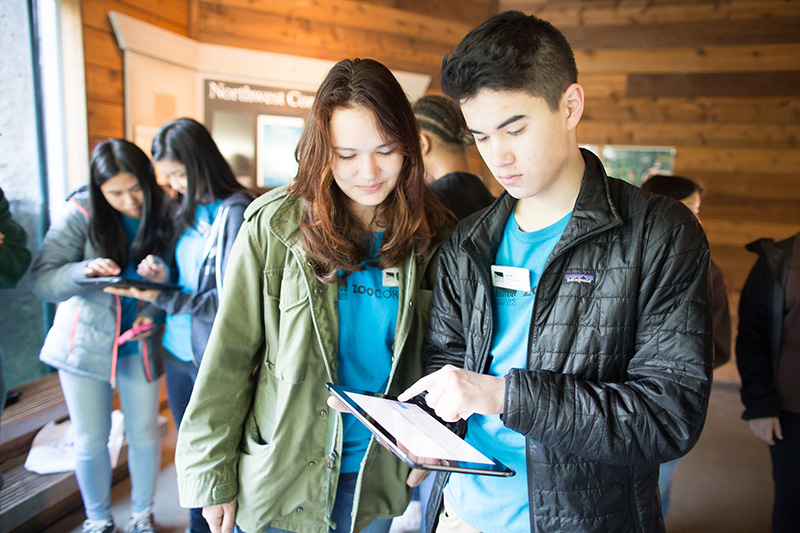 A group of young people indoors, two in the front looking at and using a tablet together, while others in the background are also working with tablets. They are wearing blue shirts and jackets.
