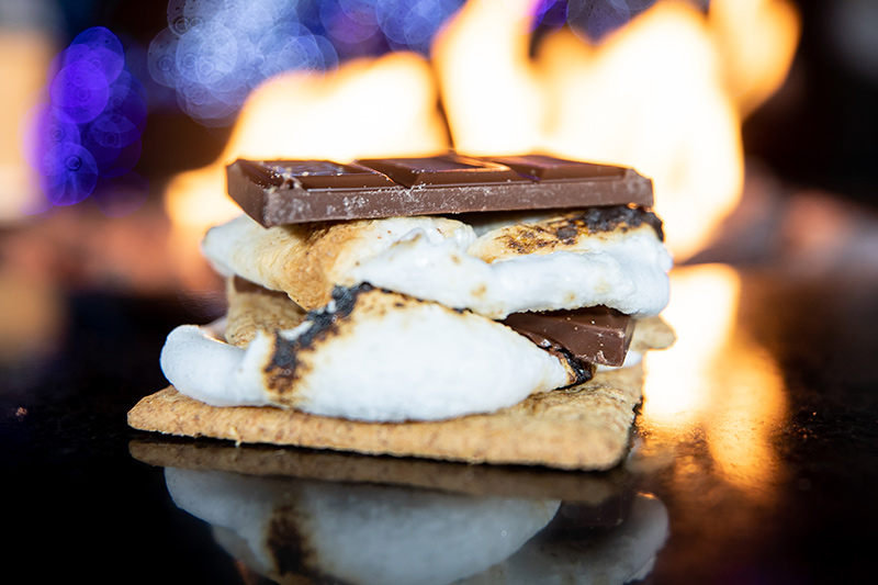 A close-up of a s’more made with toasted marshmallows, a chocolate bar, and graham crackers, with a fire burning in the blurry background.