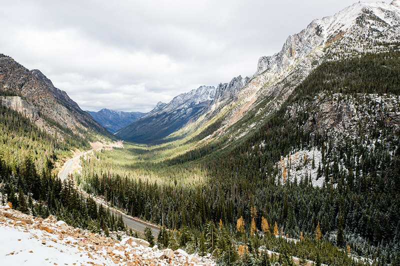 Snow-dusted mountains rise above a dense evergreen forest, with a winding road cutting through the valley below under a cloudy sky. Sunlight highlights sections of the landscape.
