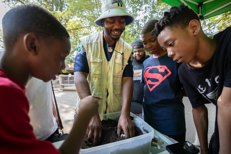 A group of boys gather around a smiling man who is teaching them about soil and plants, with their hands in a clear bin of dirt, outdoors under the shade of trees.