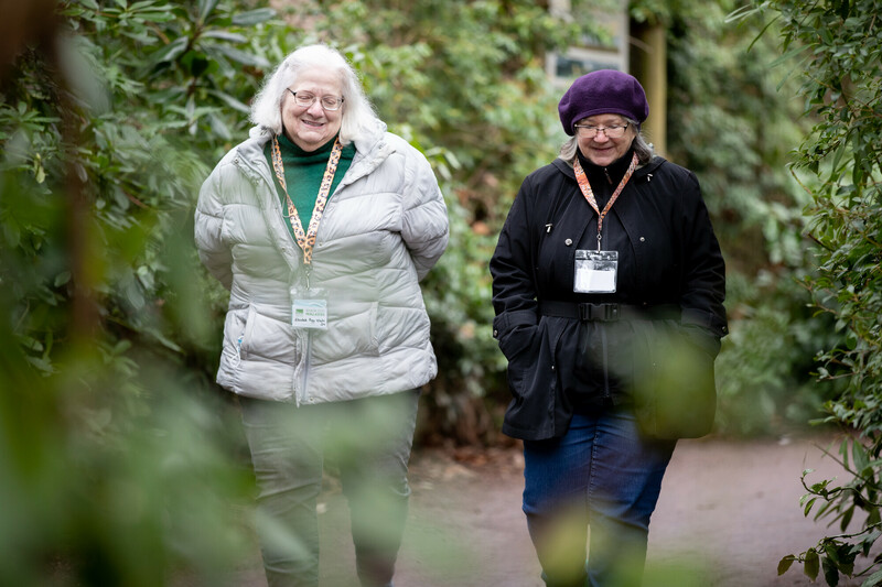 Two older women wearing jackets and name badges walk along a garden path, smiling and talking. Lush green foliage surrounds them, creating a peaceful outdoor setting.