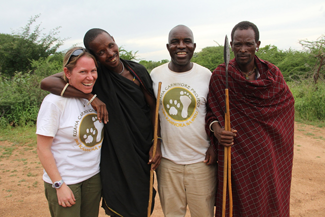Four people smile and pose together outdoors. Three men wear traditional Maasai clothing and one woman wears sunglasses and a Ruaha Carnivore Project t-shirt. Two men hold wooden sticks. Greenery is visible in the background.
