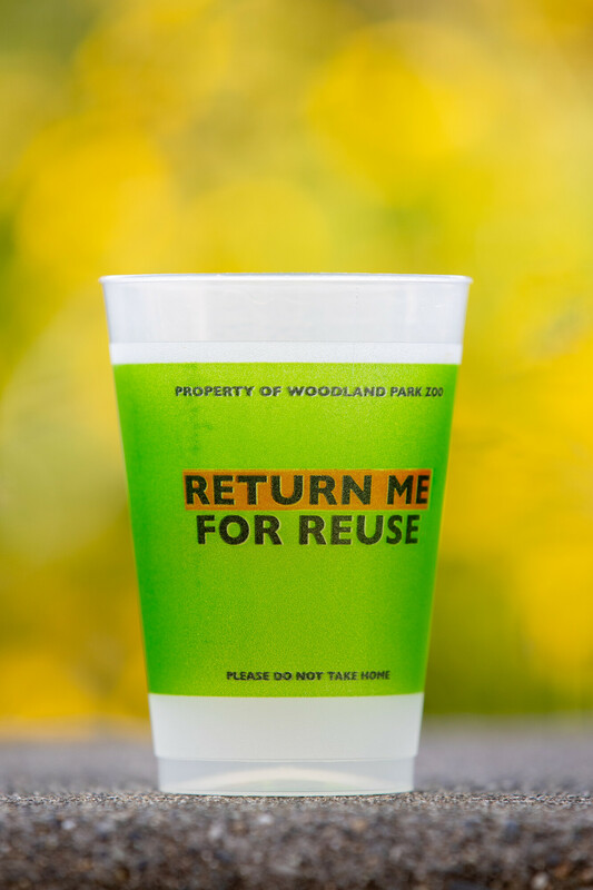 A plastic cup with a green sleeve labeled RETURN ME FOR REUSE stands on a surface, with a blurred yellow background. The cup is marked Property of Woodland Park Zoo and Please do not take home.