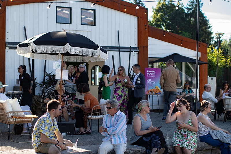 People gather outside a white barn-style building at a sunny outdoor event. Some sit and chat on chairs and benches, while others stand and mingle near umbrellas. Trees are visible in the background.