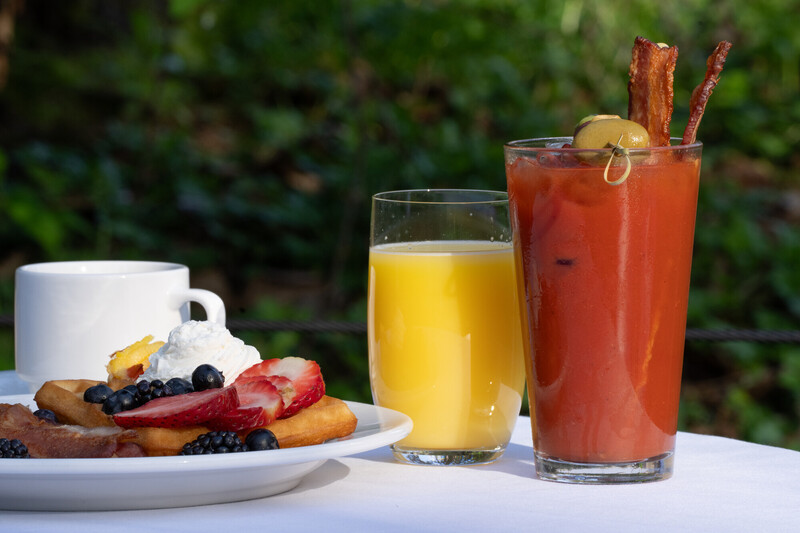 A breakfast setting with a plate of waffles topped with berries and whipped cream, a cup of coffee, a glass of orange juice, and a Bloody Mary garnished with bacon and an olive, on a white tablecloth outdoors.