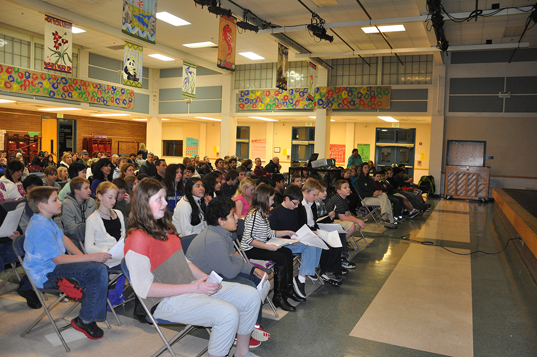 A large group of students sits in rows of chairs in a school gym or auditorium, attentively facing a stage. Colorful banners and artwork decorate the walls above them. Some students hold papers in their laps.