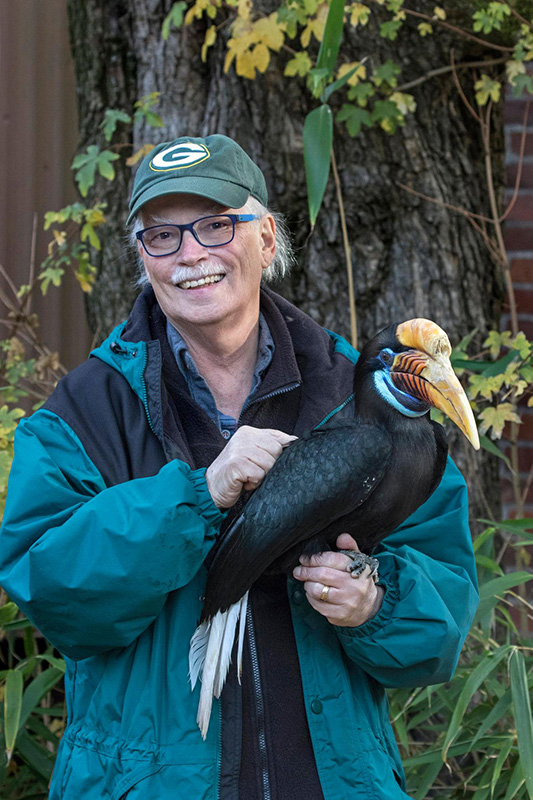 A smiling man in a green jacket and cap stands outdoors, holding a colorful hornbill bird with a large yellow-orange bill and blue markings, in front of a tree and foliage.