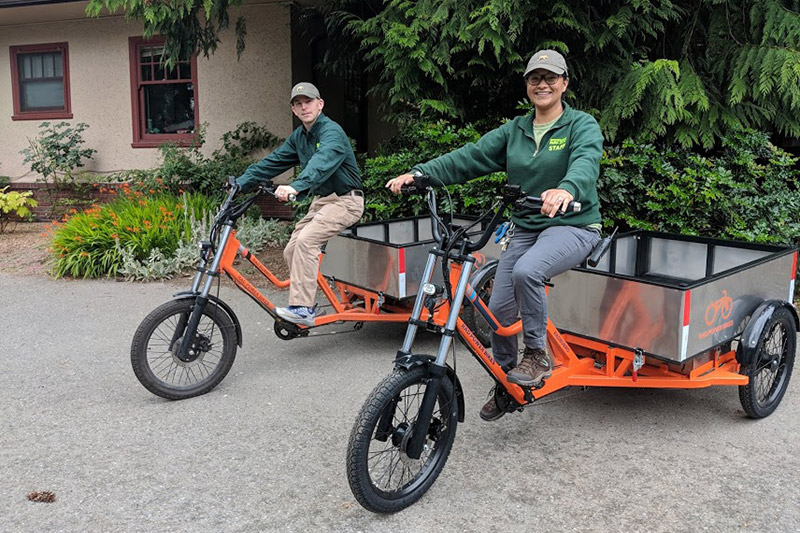Two people wearing green jackets and hats sit on large orange electric cargo tricycles with metal bins, parked on a paved area near a building and greenery.