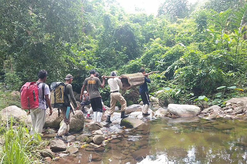 Six people carry a large wooden log across a shallow, rocky stream in a lush, green forest. Some have backpacks and plastic coverings on their legs, and the group appears to be working together to cross the water.