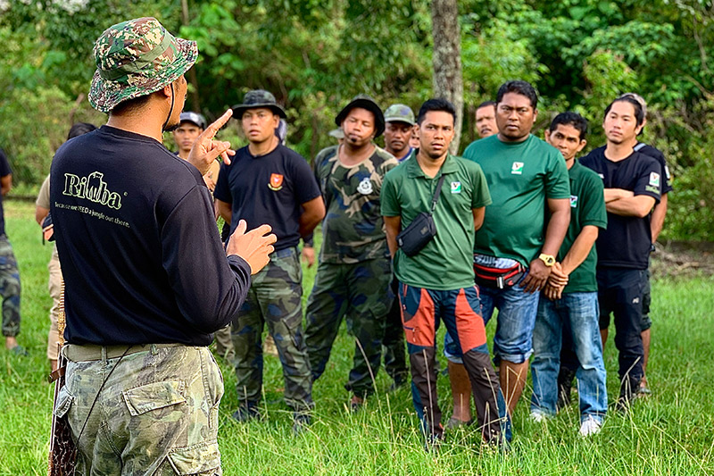 A person in camouflage addresses a group of people standing on grass in an outdoor, forested area; the group wears dark green and black attire, appearing attentive to the speaker.